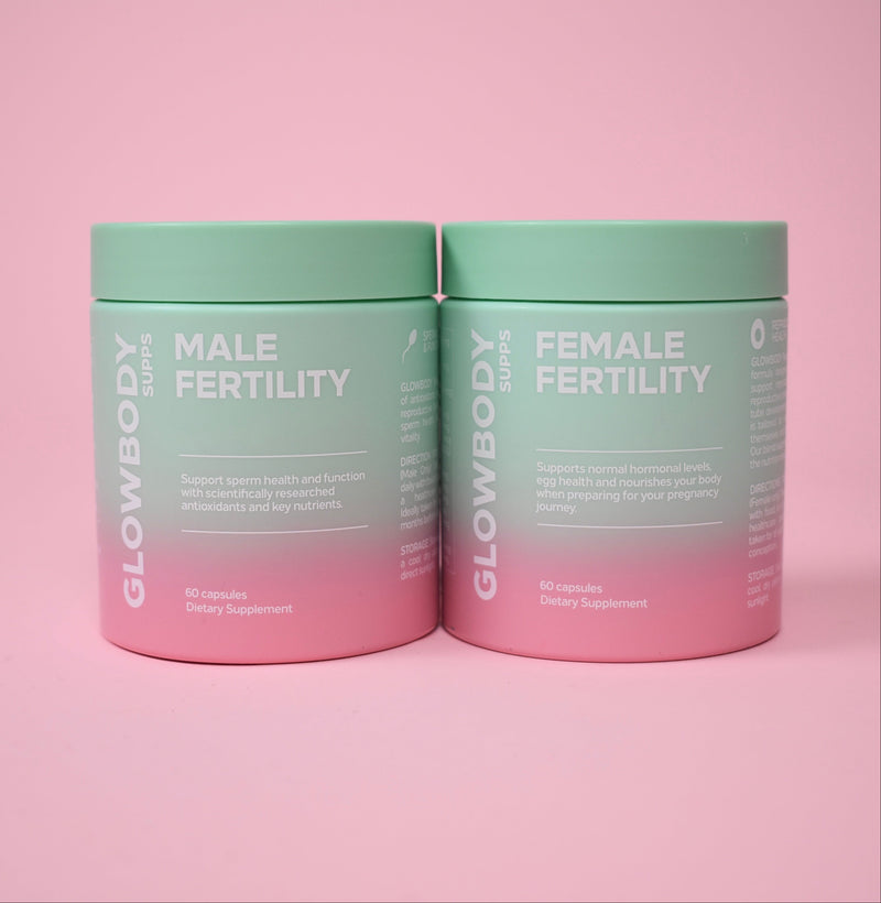 Two cylindrical supplement jars on a solid pink background. The left jar is labeled 'GLOWBODY SUPPS MALE FERTILITY' and the right jar is labeled 'GLOWBODY SUPPS FEMALE FERTILITY'.